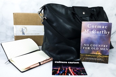 An open, blank journal with a pen, a large black bag, the book No Country for Old Men, and a card that says Culture Carton.