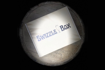A showcase of the beautiful Swizzle Box box sitting on top of a old rum barrel.  The Box has the Swizzle Box logo and palm tree shadows.
