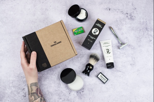 A hand hold a closed Shaving Club subscription box with a razor and other shaving products around it.