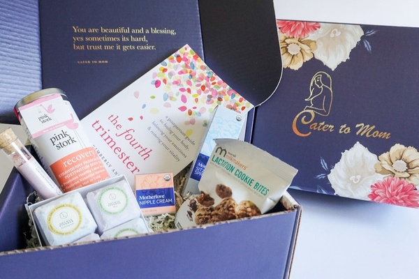 A blue and floral Cater to Mom subscription box next to an open box containing a book, tea, cookies, nipple cream and more.