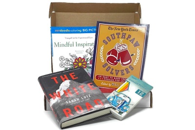 An open Coloring and Classics subscription box filled with a novel, a coloring book, a word puzzle book and colored pencils.