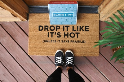 A person standing on a doormat that says Drop It Like It's Hot (Unless It Says Fragile) and there's a Smartass and Sass box.