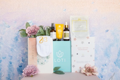 A Loti Wellness Self-Care subscription box surrounded by a Goal Getter workbook, a fragrance spray and lotion.