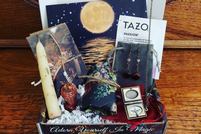 A Witchy One subscription box filled with earrings on a grey pouch, a candle, a necklace, a card and envelope and more.