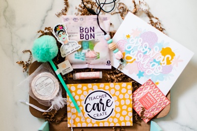 An open Teacher Care Crate subscription box filled with bon bons, an encouraging card, lip balm and a pom pom pen.