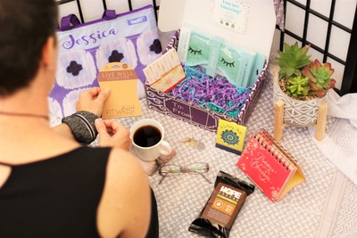 A woman looking at her Fit Wrist, sitting next to a subscription box containing a sleep mask, a protein bar and more.