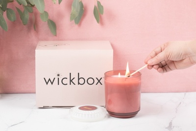 A hand using a match to light a candle in a pink jar, next to a closed Wickbox subscription box.