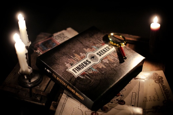 A Finders Seekers Escape Room Game subscription box with a magnifying glass on top and lit candles nearby.