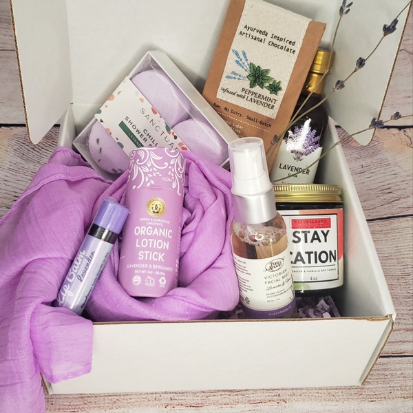 An open subscription box containing a lavender colored scarf, organic lotion stick, shower bombs and lavender body spray.
