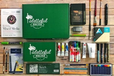 A green Paletteful Pack subscription box surrounded by art supplies including paint, brushes, markers and paper.