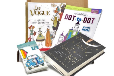 An open Coloring and Classics subscription box with a novel, a coloring book, a connect the dot book and colored pencils.
