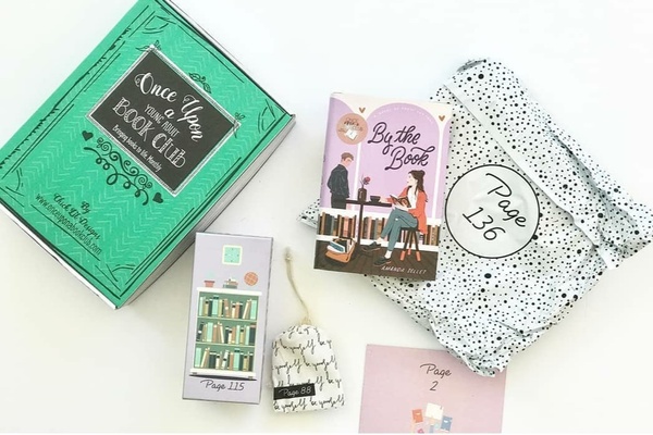 A green Once Upon a Book Club subscription box lies next to a novel along with 4 wrapped gifts with page numbers.