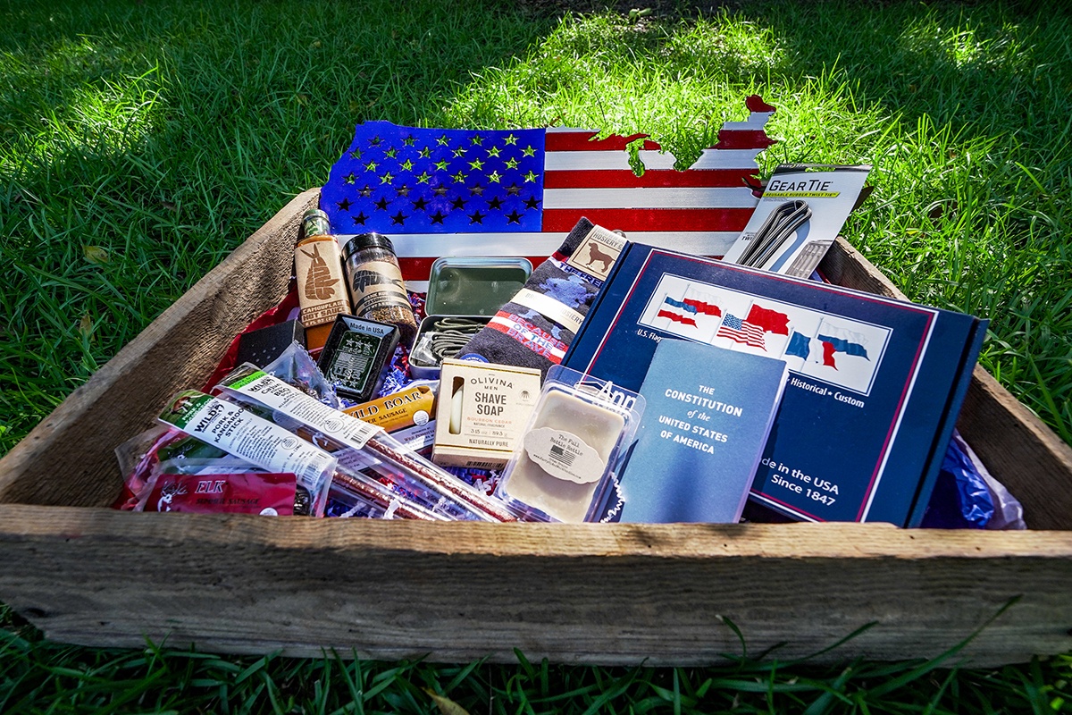 A shallow wooden box filled with items from an America's Crate subscription box including food, soap, books and more.