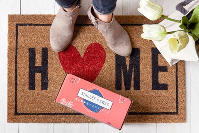 A woman standing on a welcome mat with a SinglesSwag subscription box on it.