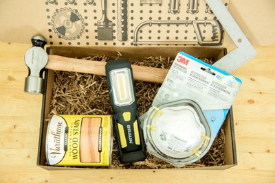 A Standard Tool subscription box filled with wood stain, a hammer, a filter mask, a hand square and a level.