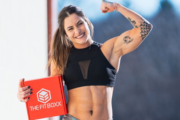 A strong woman flexing her arm and holding an orange subscription box labeled The Fit Boxx.