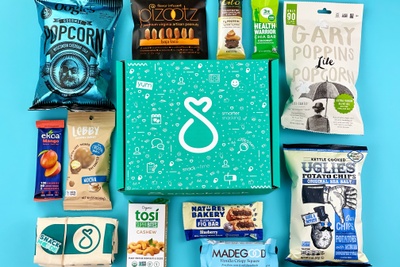 A closed, gluten free, SnackSack subscription box surrounded by popcorn, mochi, potato chips, fig bars and other snacks.