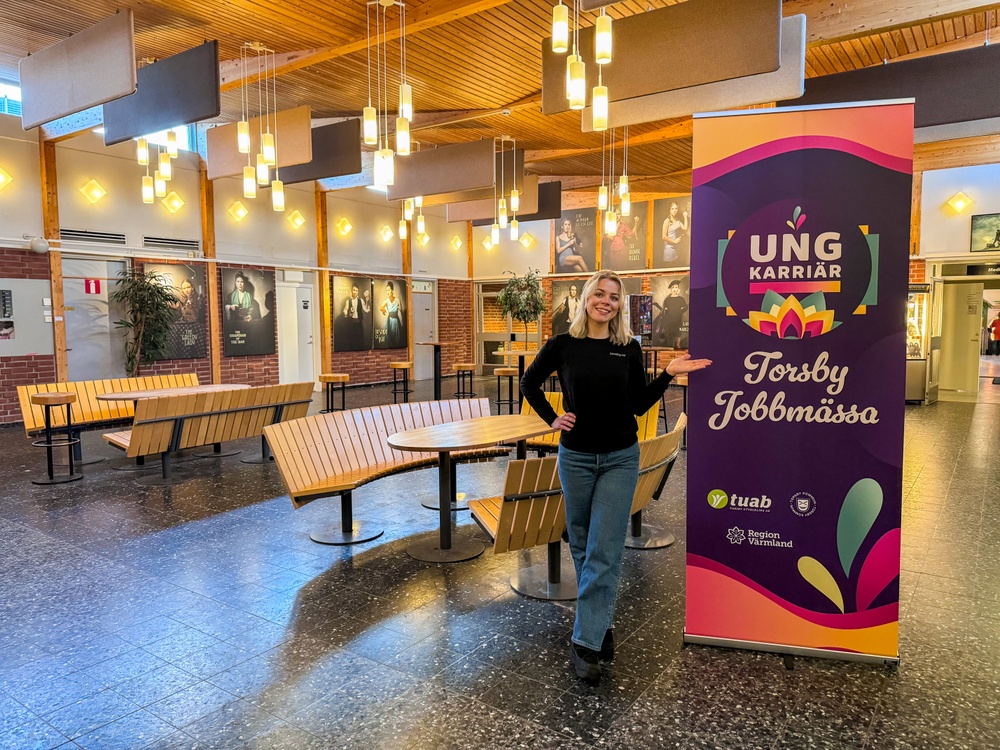 Torget på Stjerneskolan med sittplatser. En färgglad rollup med Ung Karriär - Torsby Jobbmässas logotype. Elvira Berglund, projektledare, poserar till vänster om rollupen. 