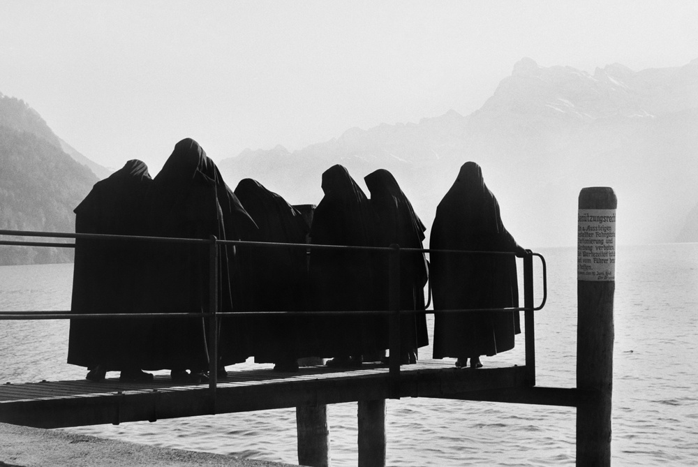 Nunnor står på en brygga vid en sjö. Fotografi av Christer Strömholm taget i Schweiz 1950