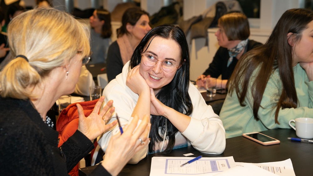 Ukrainska kvinnor sitter tillsammans med etablerade svenska kvinnor i en lokal och samtalar. De ser glada och engagerade ut. 