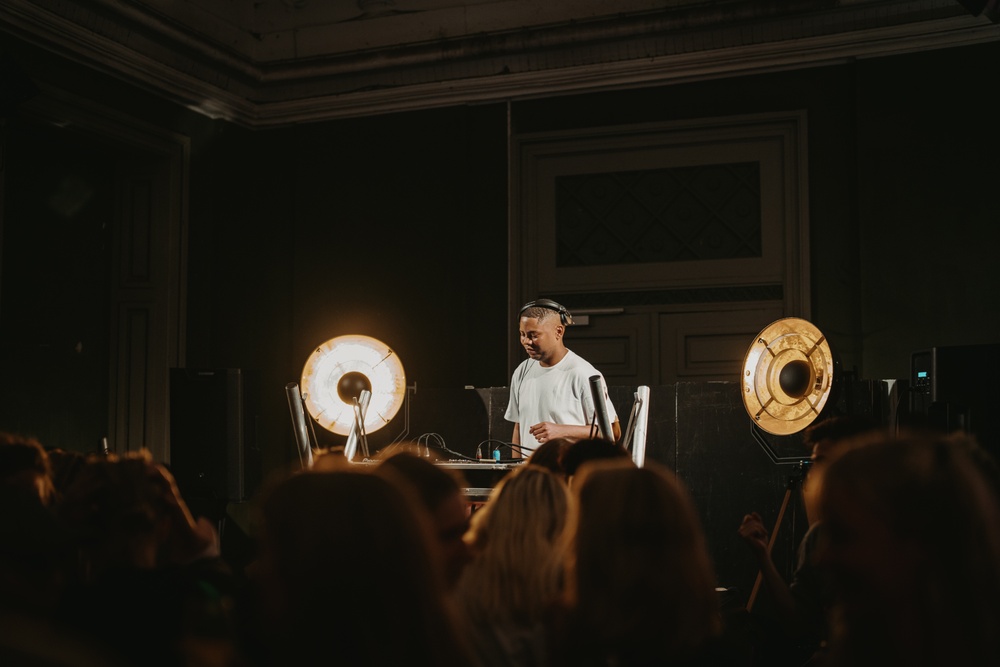 A man performing an electronic music act on a stge with two spotlights rught and left and an crowd of on-lookers.