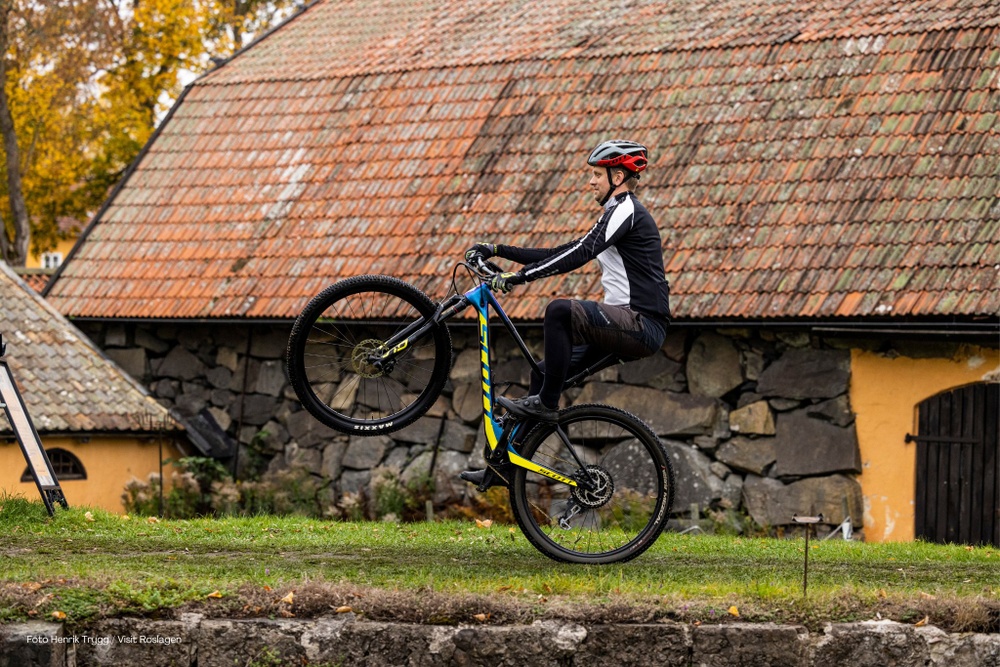 Cyklist på bakhjulen på en MTB framför en gammal gul byggnad med stenvägg och rött tegeltak. 