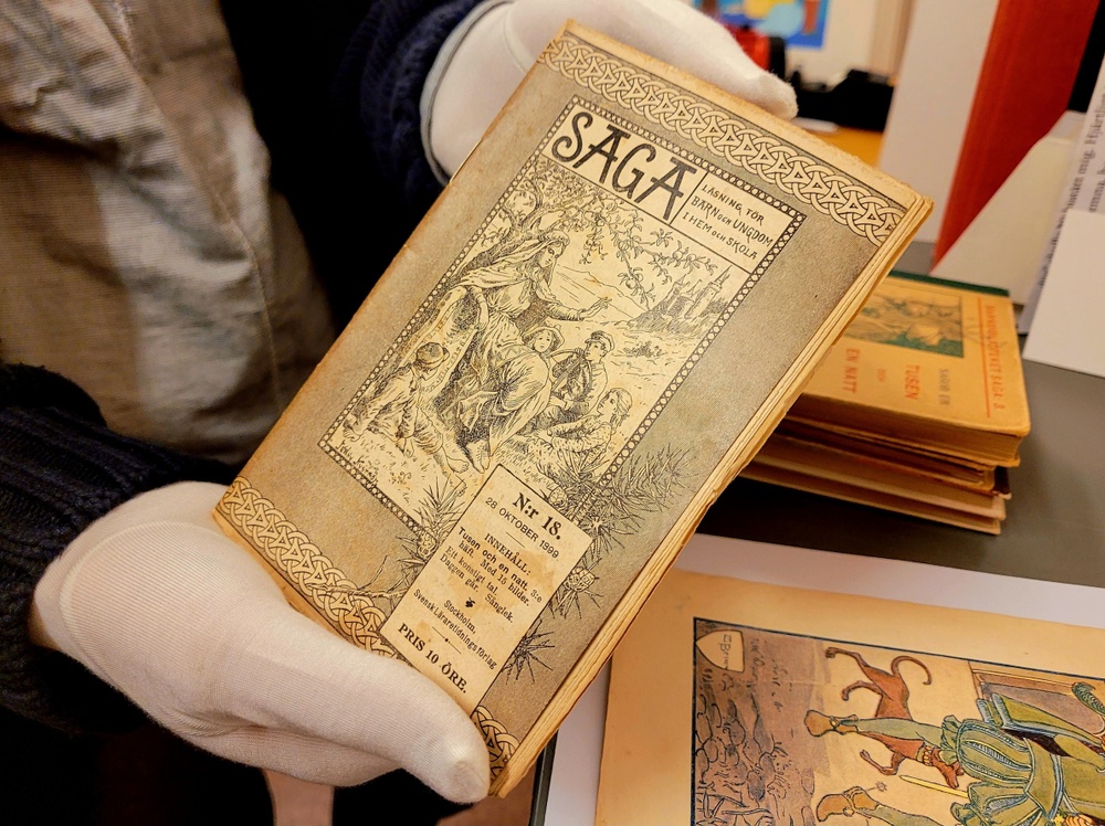En bok ur barnboksförlaget Sagas arkiv hålls av händer med arkivvantar.