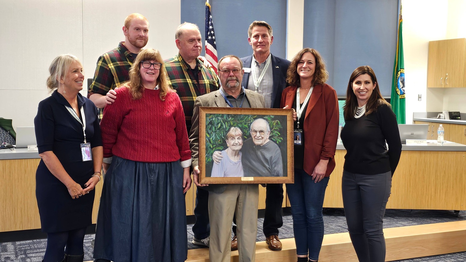 Group photo featuring members of the Peninsula School District Board and community members during the presentation of a painting of Hugh and Janice McMillan. The painting, held by one of the attendees in the center, depicts the McMillans, long-time supporters of the district. The group stands in front of an American flag and Washington State flag, smiling together to commemorate the generous donation.