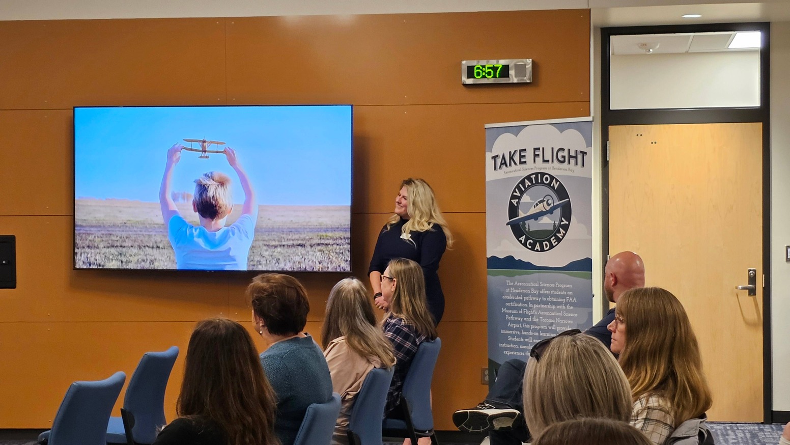 Image of a presentation at a school board meeting. The speaker, Kelsey Parke, stands beside a screen displaying a child holding a toy airplane, symbolizing the newly announced Aviation Academy at Henderson Bay High School. Next to her is a large banner with the words 'Take Flight' and the Aviation Academy logo. The audience is seated, watching the presentation.