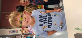 Kindergarten student with green rimmed glasses smiling and wearing a shit with text that reads "I am the future" and variety of stem icons."