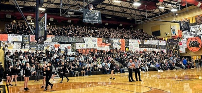 A basketball game is underway, with a packed crowd cheering on their team. The bleachers are filled with fans holding signs and banners.