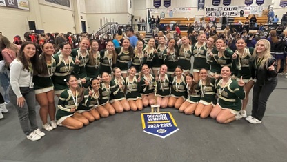 A group of high school cheerleaders sit in a half-circle by a banner that says "Division Winner 2024-2025"