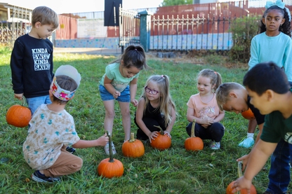 A group of kindergarteners sit on the grass, each holding a round, orange pumpkins