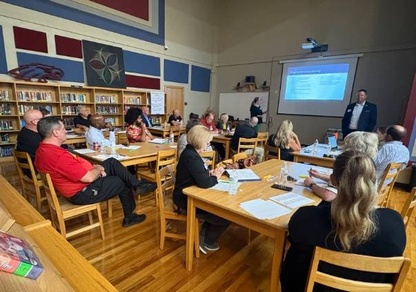 A library full of adult guests are seated at tables looking at the front of the room where two suited administrators or leading a presentation.