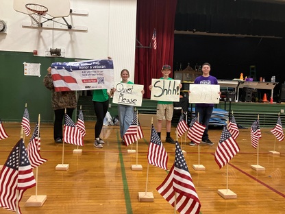 Students stand in the school gym holding signs that say "quiet please" and "shhh." Around them are small American flags.