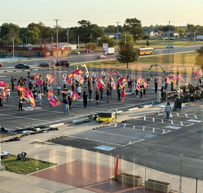 JHS Band students practice their marching show on a black asphalt-covered lot lined with yard lines. Students are twirling flags, playing instruments, and carrying equipment as the sun rises in the eastern sky. 20th street can be seen in the background and cars pass the new practice field as JHS students come to school for the day.