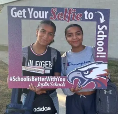 Two female JHS students stand on the sidewalk outside Kaminsky Gym in the morning sunlight. They are posed together holding a cutout frame through which their smiling faces peek. The girls hold the Eagle-branded frame together, which reads "Get Your Selfie to School" and bears the hashtag, "School Is Better With You".