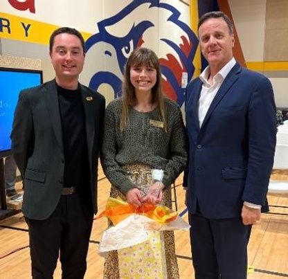 Two school leaders pose with a female high school student, smiling and holding a gift bag at a board of education meeting.