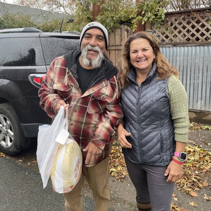 A man and woman smile at the camera. The man is holding a grocery bag with a turkey and other Thanksgiving food inside.