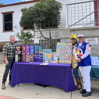 3 people stand around a purple table, which is holding various educational materials regarding substance abuse.