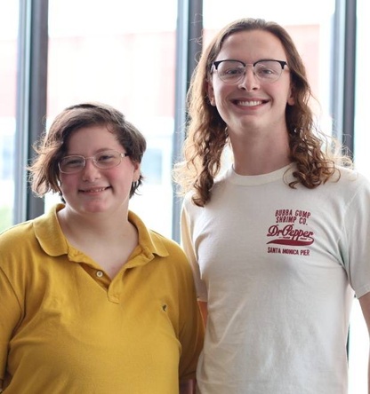 Two teenagers pose together before a brightly-lit window. Both are smiling, wearing glasses. One wears a white t-shirt, the other wears a bright yellow polo.