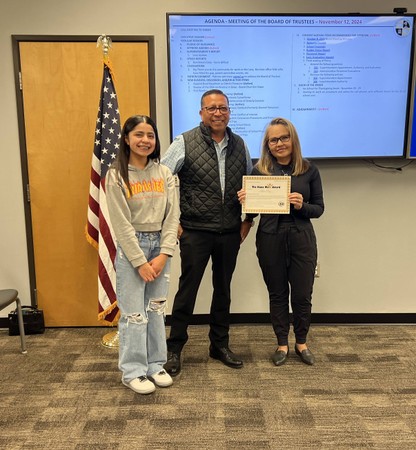 Three people standing before a flag and a display in the board room. One is holding a certificate. 