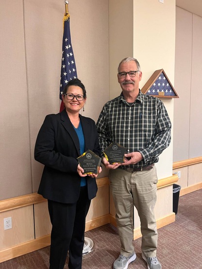 Board President Julia Burns and Board Clerk Jim Whitaker stand together in the board chambers, each holding an engraved star-shaped trophy.