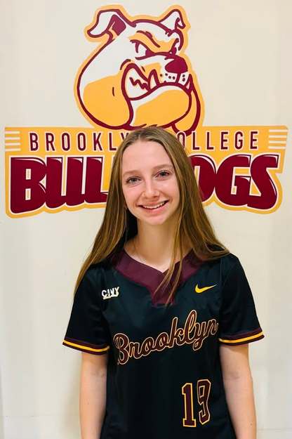 A young woman smiles for the camera, wearing a black and maroon baseball jersey with the word 'Brooklyn' and the number '19' on it. She is standing in front of a wall with a bulldog mascot logo.