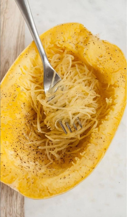 A spaghetti squash sitting on a marble and wood countertop. The squash is roasted and sprinkled with pepper. A fork sits in the squash covered in loose squash fibers.