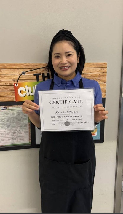 A photo of Kana, who was awarded the Sodexo Experience Award in October. She is standing next against the wall in her uniform holding up an award certificate. 