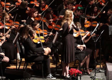 A young woman plays the flute during an orchestra performance, surrounded by violinists.