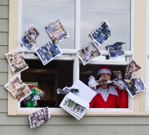 A woman dressed as an elf looks out a window, surrounded by colorful papers.