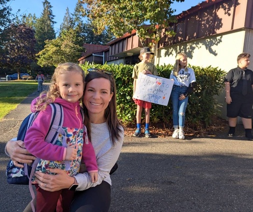 A young girl with a backpack smiles as she stands beside an adult, likely her parent, who is kneeling and smiling at the camera. In the background, older students hold a welcome sign that reads "Happy to see you TK!" They stand outside a school building on a sunny day, surrounded by greenery, creating a warm and welcoming scene for the first day of Transitional Kindergarten.