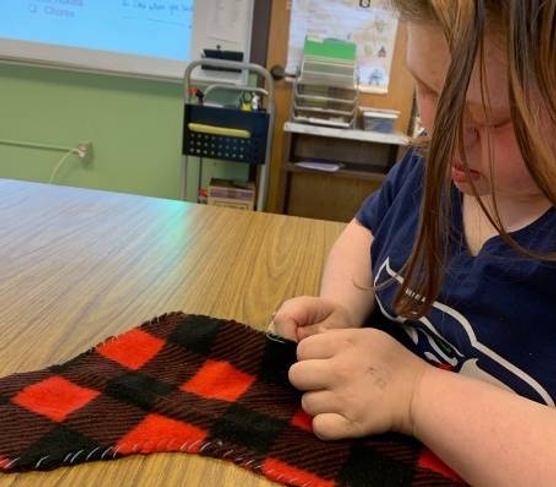 A student sits at a table, carefully sewing a red and black plaid fabric.